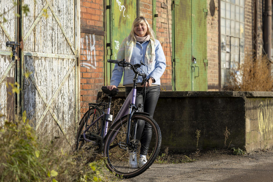 Kobieta stoi przy rowerze na tle starych, ceglastych zabudowań z dużymi, zielonymi drzwiami i zardzewiałymi elementami. Wokół widoczna jest roślinność i asfalt, a całość oświetla ciepłe światło słoneczne. 