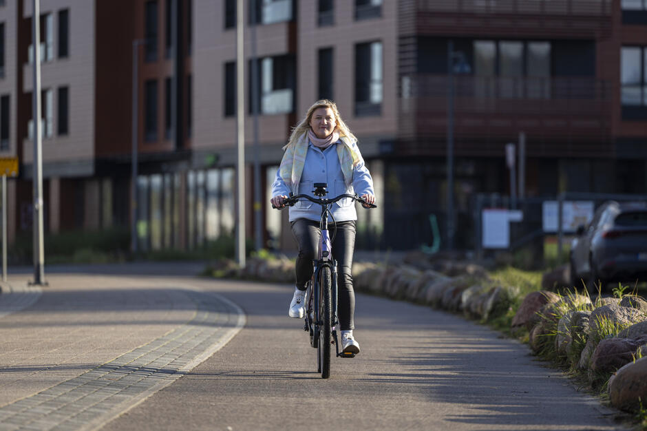 Na zdjęciu kobieta jedzie na rowerze po ścieżce rowerowej, ubrana w jasnoniebieski płaszcz i pastelowy szalik. W tle znajdują się nowoczesne budynki mieszkalne i zaparkowane samochody, a scena oświetlona jest popołudniowym słońcem.
