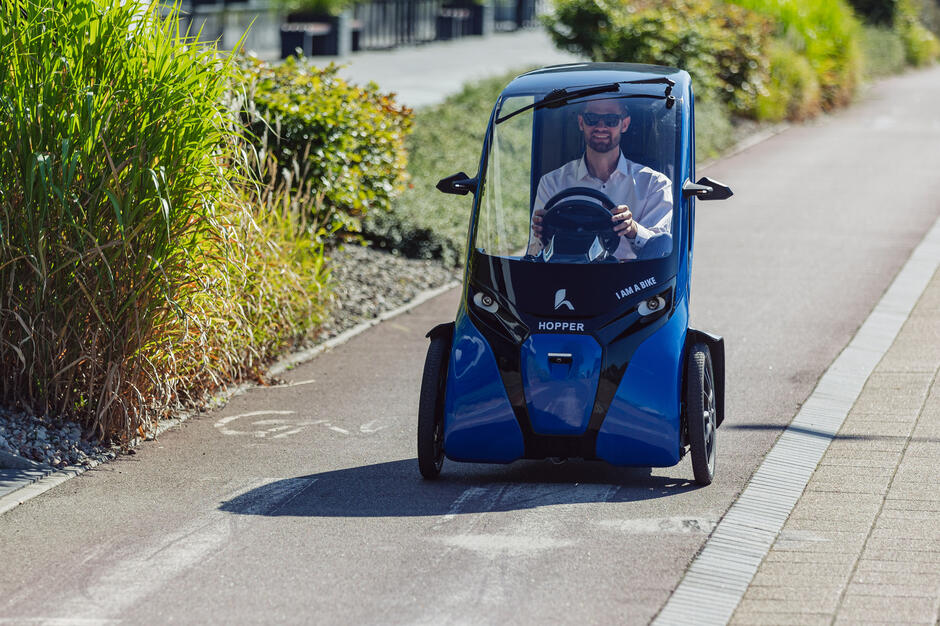 Na zdjęciu mężczyzna jedzie niewielkim pojazdem trzykołowym typu „Hopper”, przypominającym zamknięty rower elektryczny, po wyznaczonej ścieżce rowerowej. Pojazd ma niebiesko-czarną obudowę, a wokół widoczna jest wysoka roślinność i elementy miejskiej infrastruktury.

