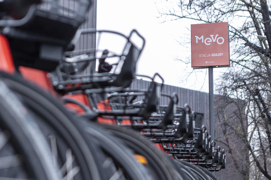 Na zdjęciu widać rząd czerwonych rowerów systemu Mevo zaparkowanych przy stacji rowerowej. W tle stoi tablica z nazwą systemu i oznaczeniem stacji „GDA267”, a po bokach widać drzewa i fragmenty miejskiej zabudowy.
