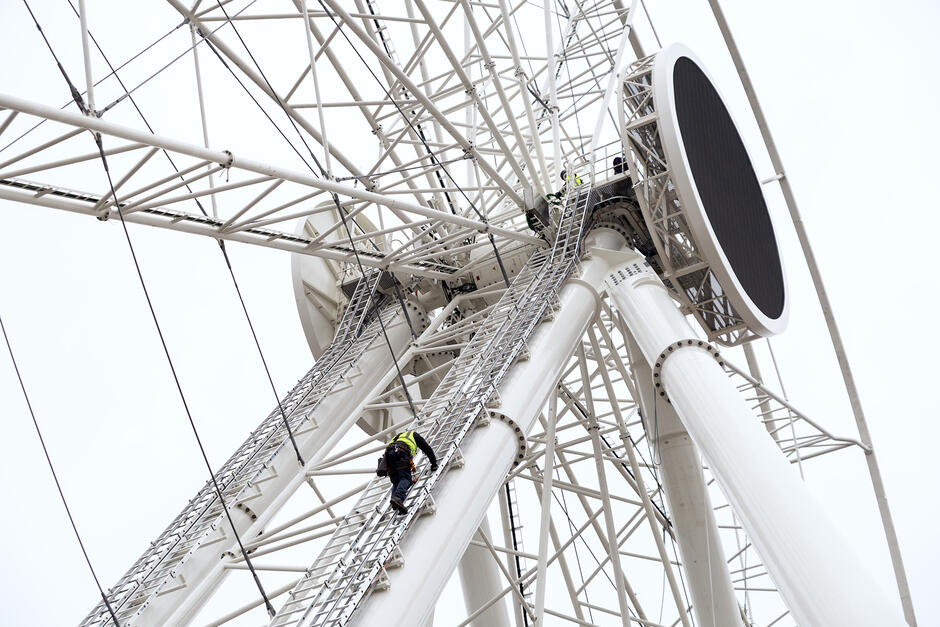 Widok fragmentu koła widokowego, na które wspina się po drabinie mężczyzna w kasku i odblaskowej zielonej kamizelce.