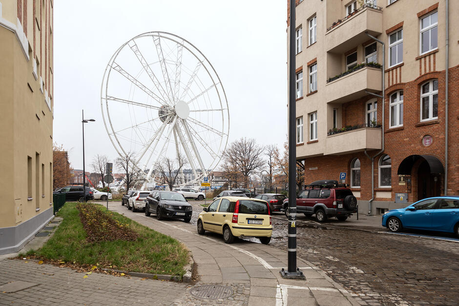 Koło widokowe w mieście widoczne między starymi domami, między którymi biegnie brukowana ulica, na której stoją zaparkowane samochody.