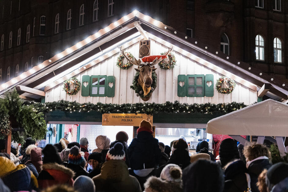 na zdjęciu chatka jarmarkowa, na górze umieszczona jest głowa łosia, przed chatką stoi tłum ludzi w zimowych ubraniach