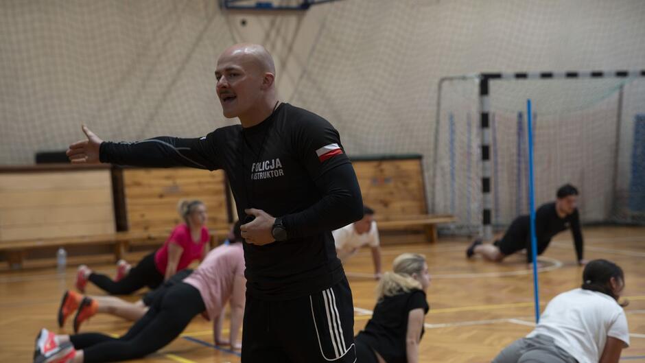 Młodzi ludzie ćwiczą na sali gimnastycznej, instruuje ich mężczyzna w dresie z napisem Policja .