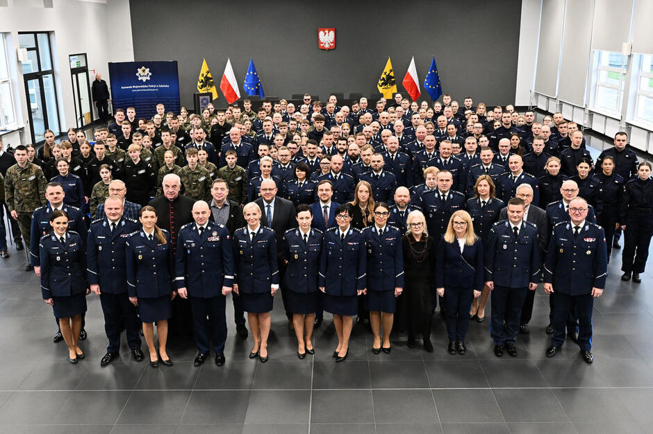 Duża grupa osób w mundurach policyjnych oraz szkół mundurowych pozuje do wspólnego zdjęcia w sali Ośrodka Szkolenia Policji.