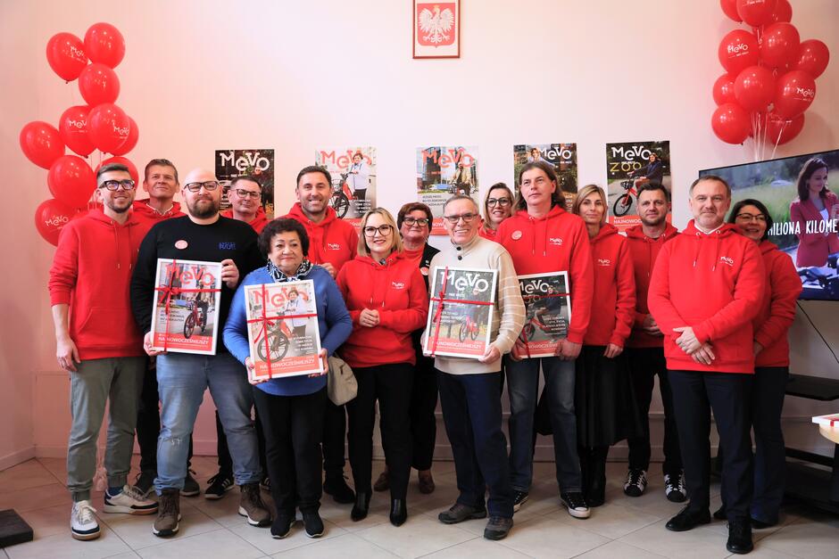 Cztery osoby trzymają oprawione w ramy plakaty ze swoimi zdjęciami na rowerach, z nimi stoją i pozują do zdjęcia osoby w czerwonych bluzach.