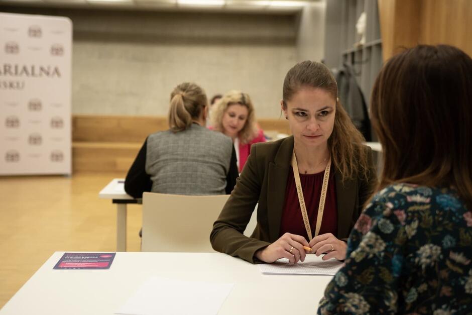 Dwie osoby siedzą przy stole, rozmawiając. Jedna jest widoczna twarzą do nas, druga - od strony pleców 