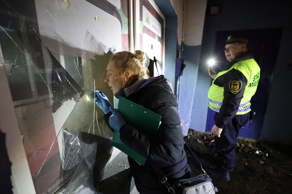 Kobieta z latarką zagląda przez popękaną szybę do tymczasowego schronienia osób bezdomnych. Obok stoi strażnik miejski i świeci w okno latarką.