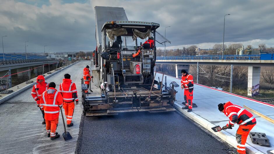na zdjęciu robotnicy w roboczych pomarańczowych kombinezonach, przed nimi jedzie duża maszyna wylewająca i wyrównująca asfalt na jezdni