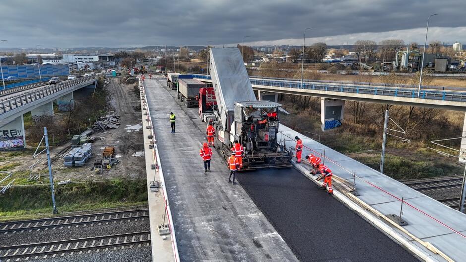 na zdjęciu robotnicy w roboczych pomarańczowych kombinezonach, przed nimi jedzie duża maszyna wylewająca i wyrównująca asfalt na jezdni