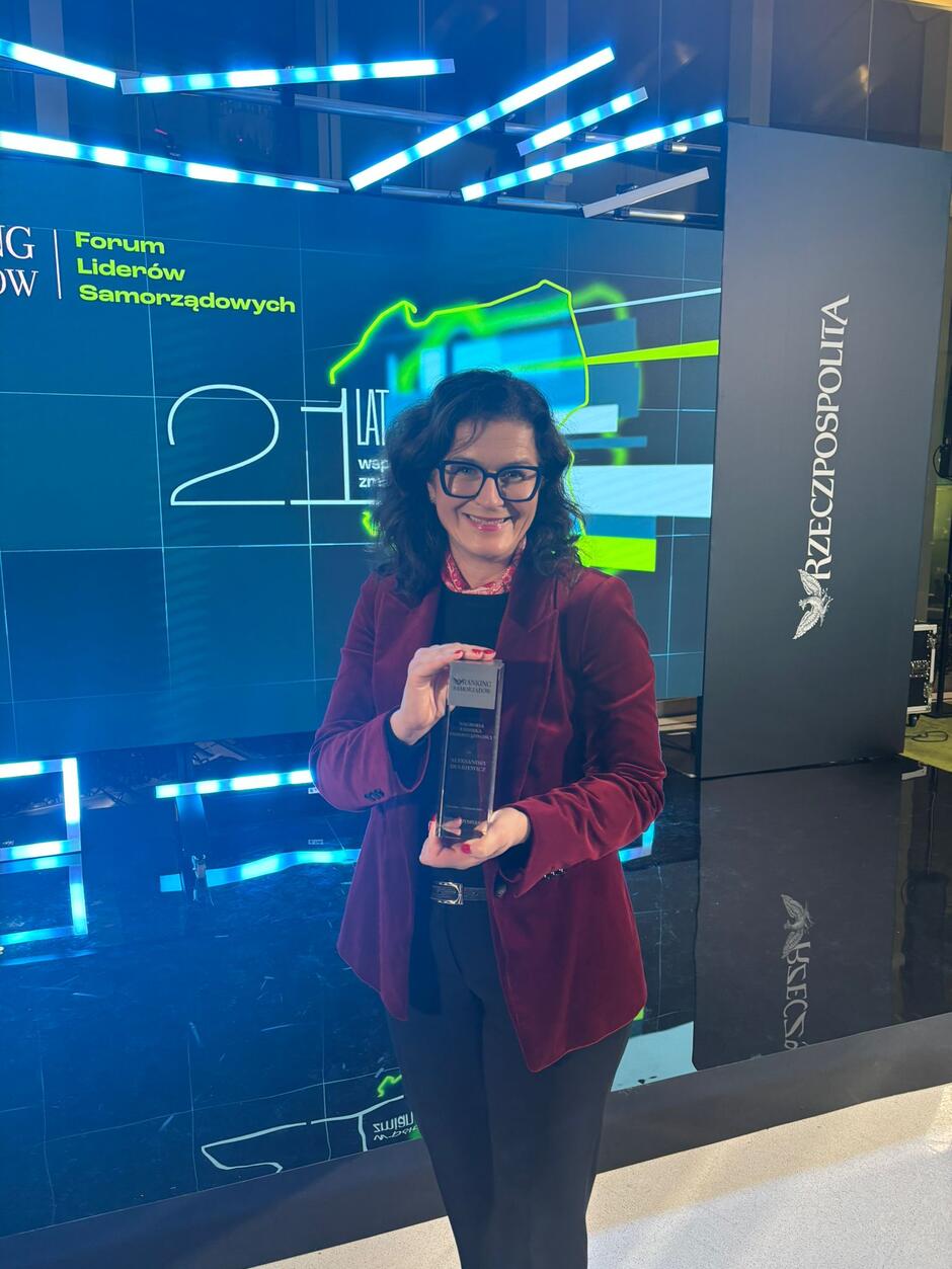 na zdjęciu kobieta w średnim wieku, w bordowej marynarce i okularach, pozuje do zdjęcia ze szklaną statuetką