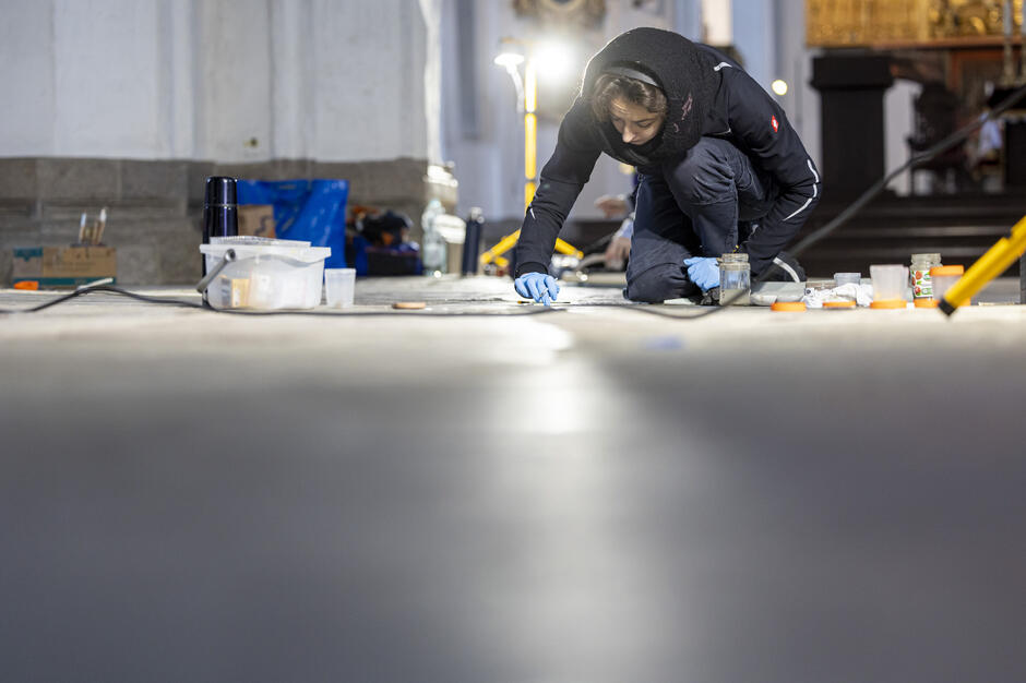 młoda kobieta, bardzo ciepło ubrana maluje coś malym pędzelkiem po kamiennej posadzce, we wnętrzu zabytkowego budynku