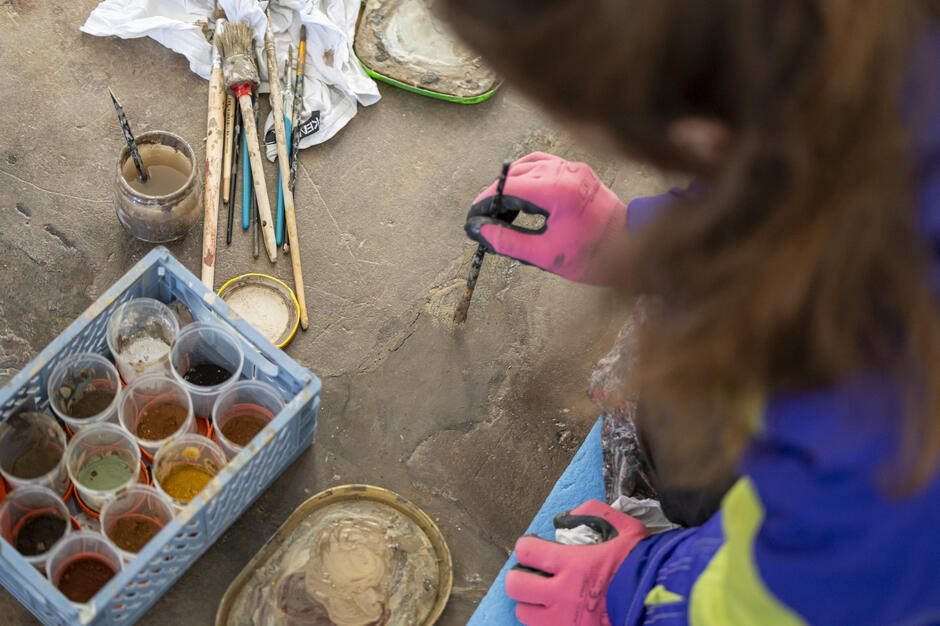 młoda kobieta, bardzo ciepło ubrana maluje coś malym pędzelkiem po kamiennej posadzce, obok widoczne różne rodzaje pędzli oraz pojemniki z farbą