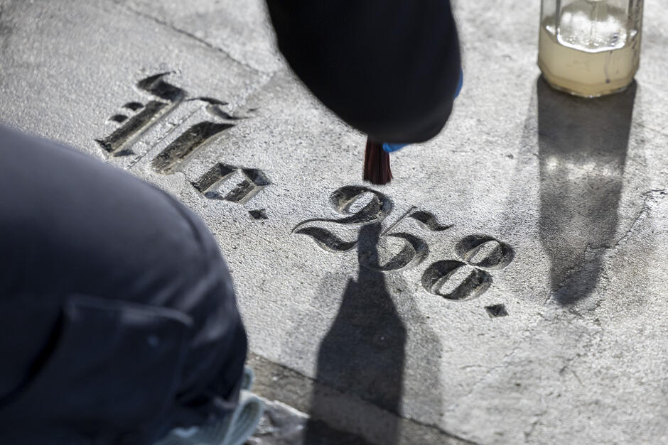  ręka osoby trzymająca szczoteczkę do malowania i pisząca na betonowej powierzchni. Osoba nosi czarną kurtkę i wydaje się być w trakcie tworzenia projektu lub wzorca na betonie. Jest mały szklany słoik z białym płynem obok osoby, który jest prawdopodobnie świecą lub szklanką wody. W środku obrazu znajduje się kilka liczb zapisanych czcionką, z numerami 1, 2, 3, 4, 5, 6, 7, 8, 9, 10, 11, 12, 13, 14, 15, 16, 17, 18, 19, 20, 21, 22, 23, 24, 25, 26, 27, 28, 29, 30, 31, 32, 33, 34, 35, 36, 37, 38, 39, 40, 41, 42, 43, 44, 45, 46, 47, 48, 49, 50, 51, 52, 53, 54, 55, 56, 57, 58, 59, 60, 61, 62, 63, 64, 65, 66, 67, 68, 69, 70, 71, 72, 73, 74, 75, 76, 77, 78, 79, 80, 81, 82, 83, 84, 85, 86, 87, 88, 89, 90, 91, 92, 93, 94, 95, 96, 97, 98, 99, i 97.