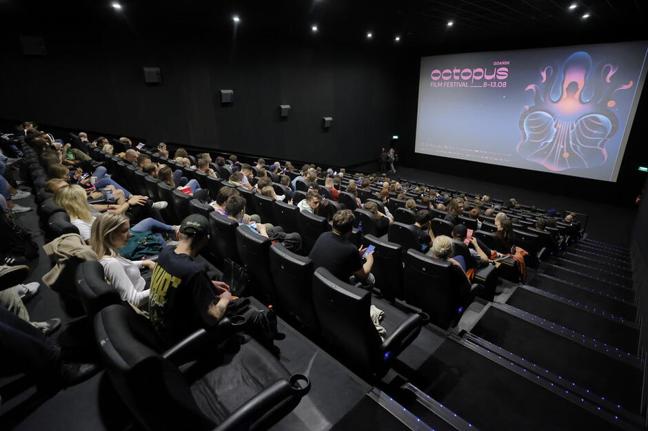 Zdjęcie wykonano podczas Octopus Film Festival. Widać salę kinową oraz siedzących na fotelach ludzi.