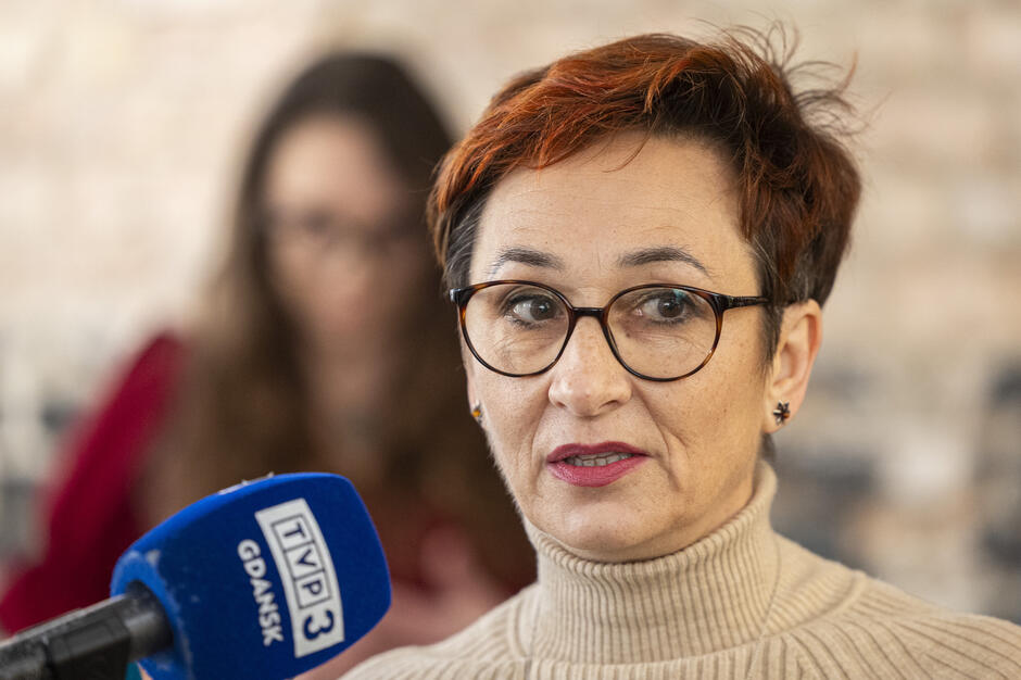  kobieta w średnim wieku z krótkimi czerwonymi włosami i okularami mówiąca do niebieskiego mikrofonu, nosi beżowy sweter z żółtawą szyją i wygląda jakby była na konferencji prasowej lub wywiadze, tło jest zamazane, ale wydaje się, że stoi przed ceglaną ścianą z inną kobietą widoczną w tle.