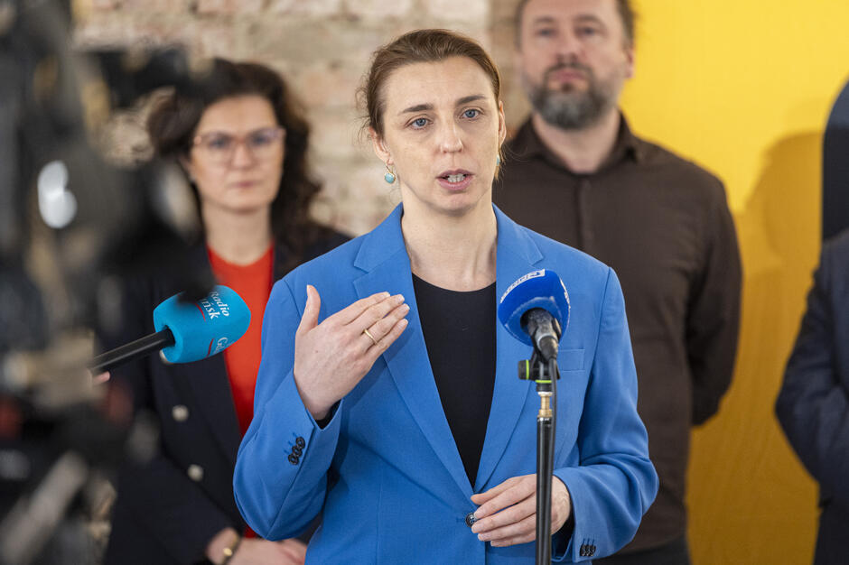  kobieta mówiąca do mikrofonu, ma na sobie niebieski blezer i wydaje się mówić z pasją, za nią są dwie inne osoby, z których jedna jest mężczyzną z brodą, tło jest rozmyte, ale wygląda na to, że kobieta stoi przed ceglaną ścianą, a do nich dołączone jest kilka mikrofonów z niebieskimi mikrofonami, sugerującymi, że wypowiada przemówienie lub składa oświadczenie.