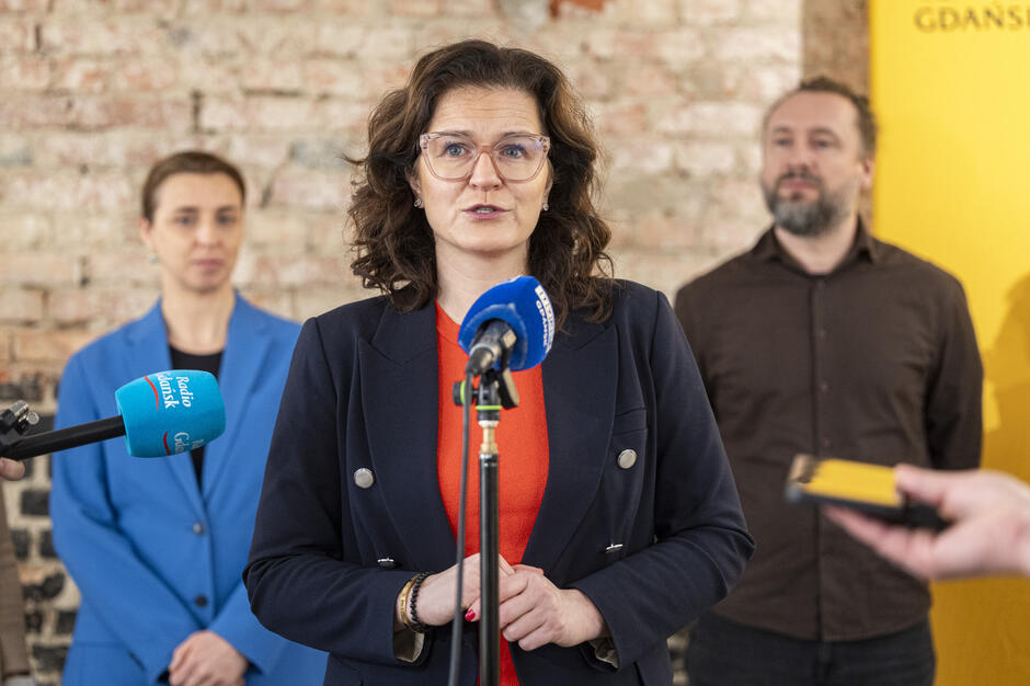  Kobieta w średnim wieku z kręconymi włosami i okularami w ramionach mówi do mikrofonu, ma na sobie czarną bluzę i czerwony top, tło jest niewyraźne, ale wygląda na to, że jest to wnętrze z ceglaną ścianą, widoczna jest głowa innej osoby w prawym dolnym rogu.