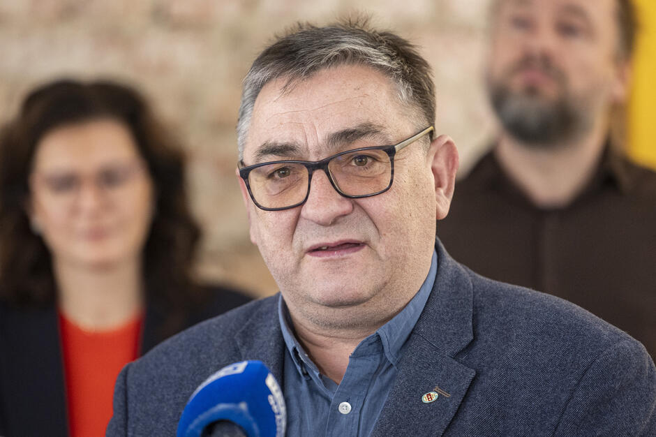  mężczyzna w średnim wieku z krótkimi siwymi włosami i okularami mówiący do mikrofonu, ma na sobie niebieski blezer i wydaje się mówić z pasją, za nim dwie inne osoby, mężczyznę i kobietę, którzy również słuchają uważnie, tło jest zamazane, ale wygląda na to, że są w pokoju z ceglaną ścianą.