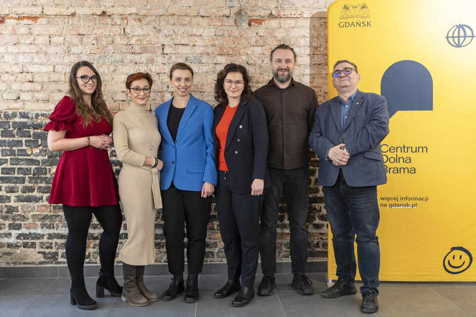  grupa siedmiu osób stojących przed żółtym banerem z logo Gdańska i tekstem Centrum Dolna Drama . Baner znajduje się w pokoju z ceglaną ścianą w tle. Ludzie stoją blisko siebie i uśmiechają się do kamery. Po lewej stronie stoi kobieta w czerwonej sukience i okularach, po prawej stronie dwóch mężczyzn w garniturach i krawatach. W środku dwie kobiety noszą niebieskie blezery, a jeden mężczyzna w pomarańczowej koszuli i czarnych spodniach. Wszystkie zdają się pozować do zdjęcia.