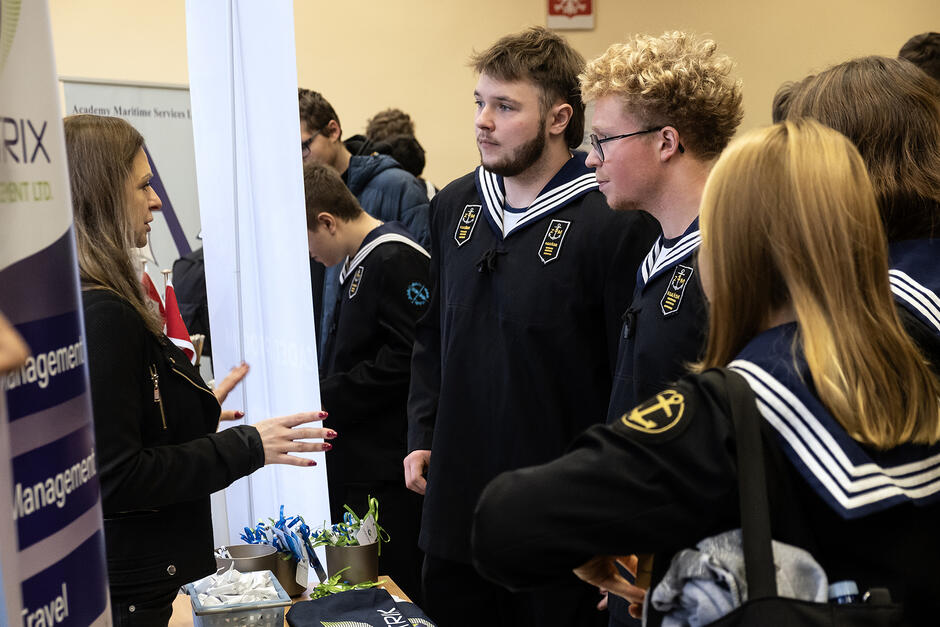 Dwóch młodych chłopaków w mundurach marynarskich stoi przed stoiskiem, na którym mówi do nich kobieta.