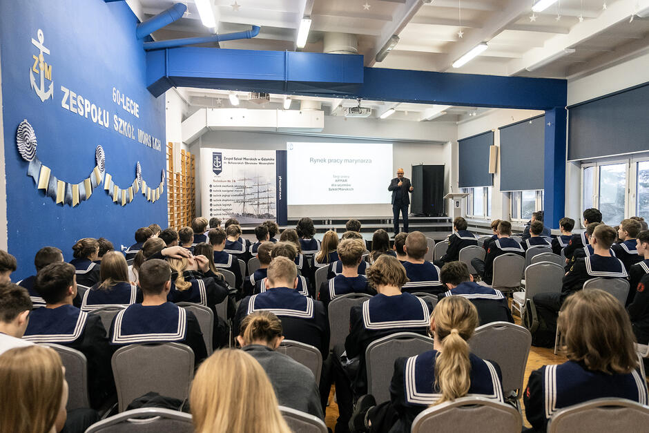 Mężczyzna przemawia w sali gimnastycznej obok ekranu z napisem Rynek pracy marynarza . Przed nim siedzą uczniowie w mundurach marynarskich.