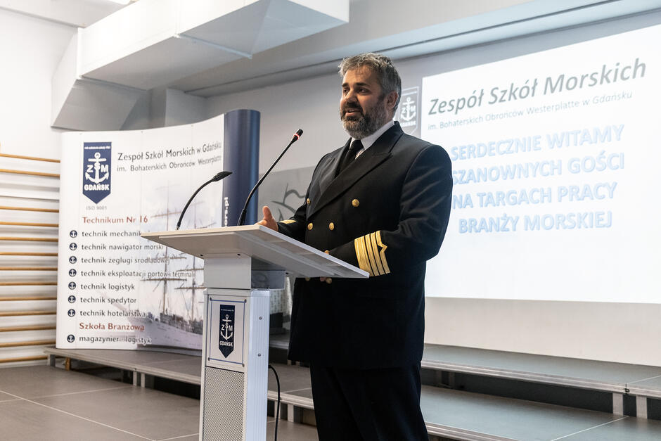Mężczyzna w mundurze marynarskim i z brodą przemawia na mównicy w sali gimnastycznej szkoły.