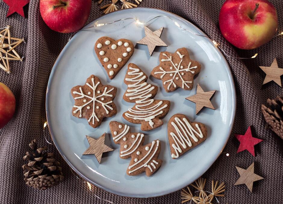 Na talerzu ułożono pierniczki w kształcie choinek, serc i śnieżynek, ozdobione białym lukrem. Wokół znajdują się drewniane gwiazdki, szyszki, jabłka i delikatne światełka, tworzące świąteczną atmosferę.
