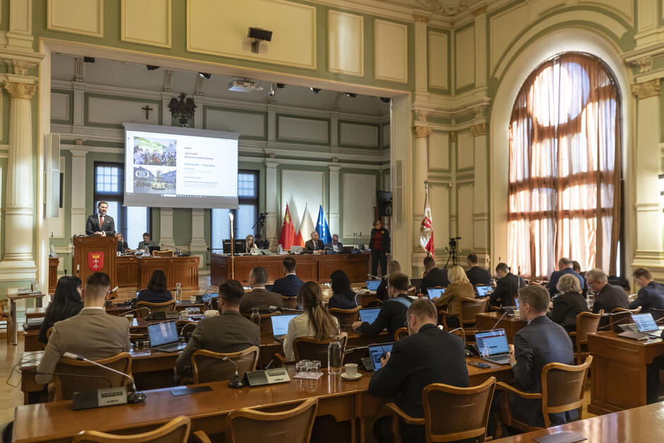 W dużej reprezentacyjnej sali siedzą radni miasta. Na mównicy przemawia mężczyzna, za nim ekran z prezentacją.