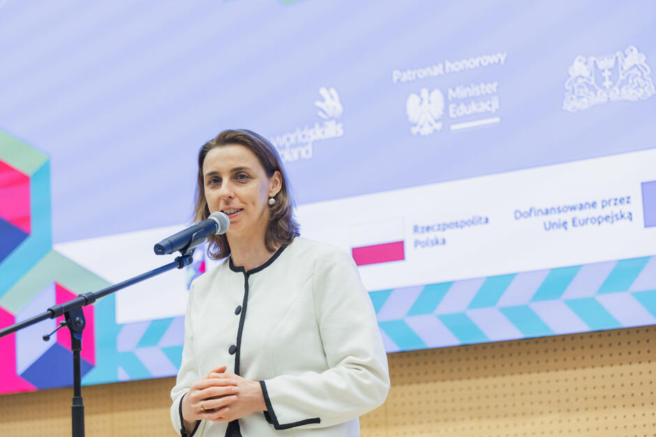 na zdjęciu kobieta w jasnej marynarce, mówi do mikrofonu