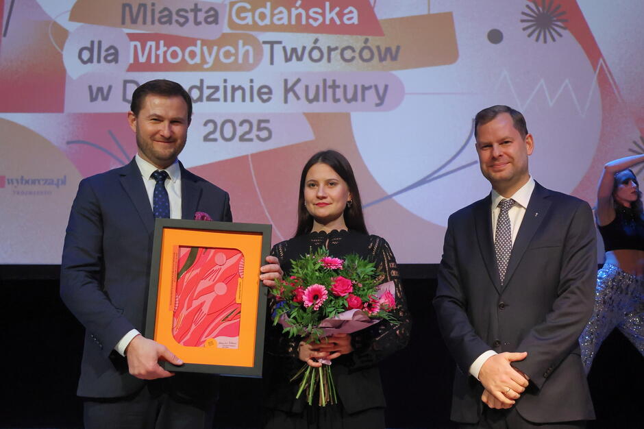 Trzy osoby pozujące do zdjęcia. Kobieta stoi pośrodku z bukietem kwiatów 