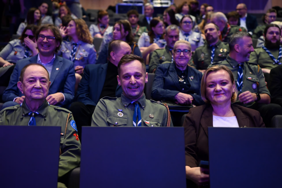 Na zdjęciu widać grupę osób siedzących na widowni podczas oficjalnego wydarzenia, w tym harcerzy w mundurach oraz osoby w eleganckich strojach. W pierwszym rzędzie trzy osoby patrzą w stronę obiektywu i uśmiechają się, a za nimi tłum uczestników koncentruje się na przebiegu spotkania.
