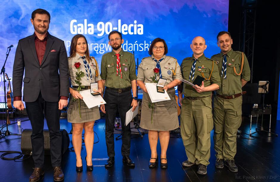 Na zdjęciu stoi sześć osób na scenie podczas uroczystej gali, a w tle widać niebieski ekran z napisem „Gala 90-lecia Chorągwi Gdańskiej”. Kilka osób w mundurach harcerskich trzyma w rękach medale, dyplomy i róże, pozując do wspólnego zdjęcia.
