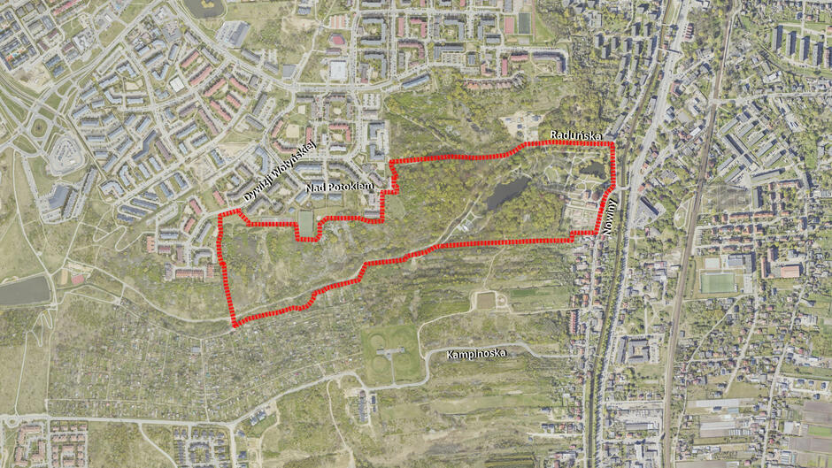 Na zdjęciu widać mapę lotniczą z zaznaczonym na czerwono obszarem o nieregularnym kształcie, obejmującym rozległy teren zielony z lasem i stawem. Wokół wyświetlonego obszaru znajdują się gęsto zabudowane osiedla oraz główne ulice.
