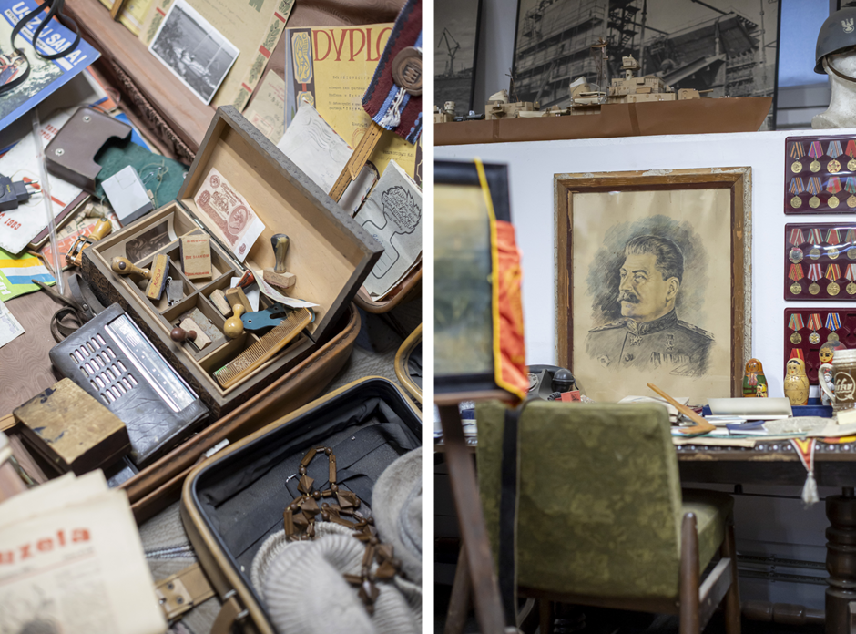Stół i walizki wypełnione są różnymi przedmiotami z minionych dekad, takimi jak dokumenty, pieczątki, banknoty, odznaczenia i drobne narzędzia. W pomieszczeniu wisi portret radzieckiego przywódcy w mundurze, otoczony kolekcją medali i pamiątek historycznych.
