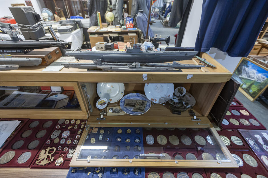 Na drewnianej gablocie stoją modele okrętów wojennych, a obok nich ustawiono drobne pamiątki marynistyczne. Wewnątrz przeszklonych szuflad znajduje się rozbudowana kolekcja medali, odznak i okolicznościowych talerzy dekoracyjnych.
