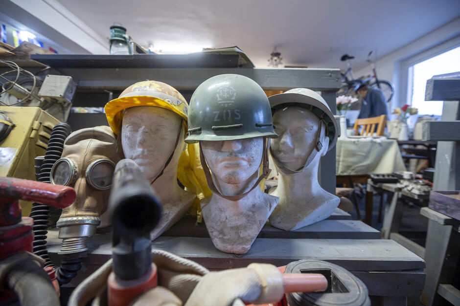 Na manekinach ustawiono różne hełmy ochronne i wojskowe, a obok nich znajduje się maska przeciwgazowa z długim wężem. Całość otacza gęsto ustawiony sprzęt techniczny i narzędzia, tworząc wrażenie magazynu dawnych wyposażeniowych pamiątek.
