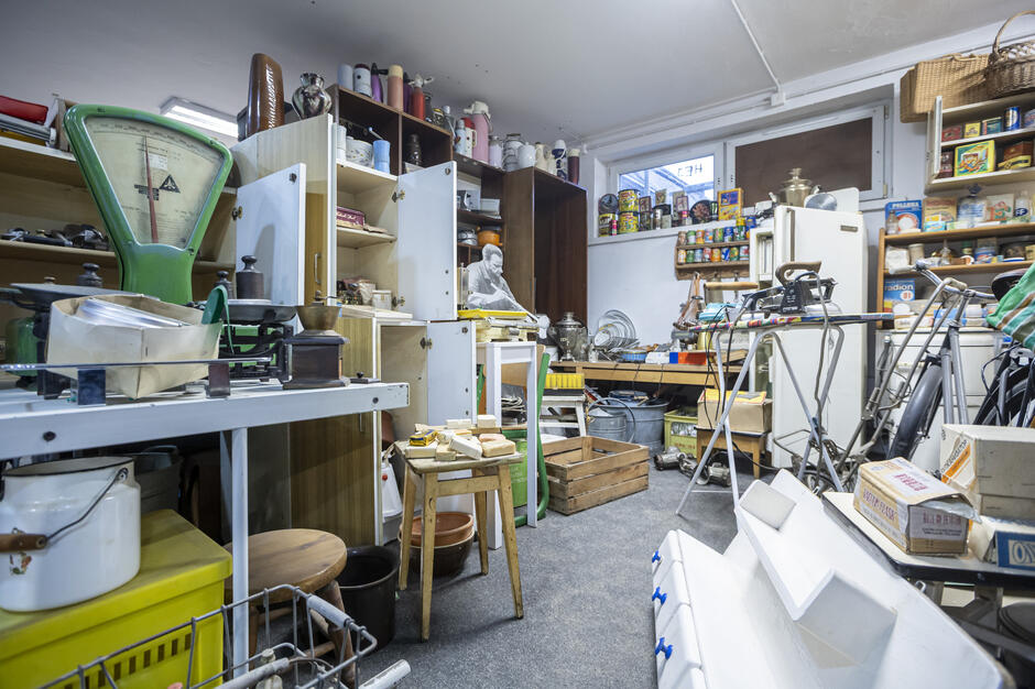 Pomieszczenie wypełniają dawne sprzęty gospodarstwa domowego, takie jak wagi, żelazka, naczynia i termosy, ustawione na półkach i stołach. W tle widać starą lodówkę, półki z opakowaniami produktów spożywczych i narzędzia, tworzące atmosferę muzealnej rekonstrukcji sklepu lub zaplecza z minionych dekad.
