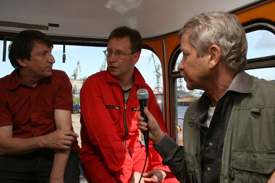 Trzech mężczyzn siedzi wewnątrz pojazdu lub kabiny, rozmawiając ze sobą, a jeden z nich trzyma mikrofon skierowany do rozmówcy w czerwonym kombinezonie. W tle przez okno widać żurawie portowe i fragment nabrzeża, co sugeruje rozmowę związaną z pracą lub wydarzeniem w stoczniowym otoczeniu. 
