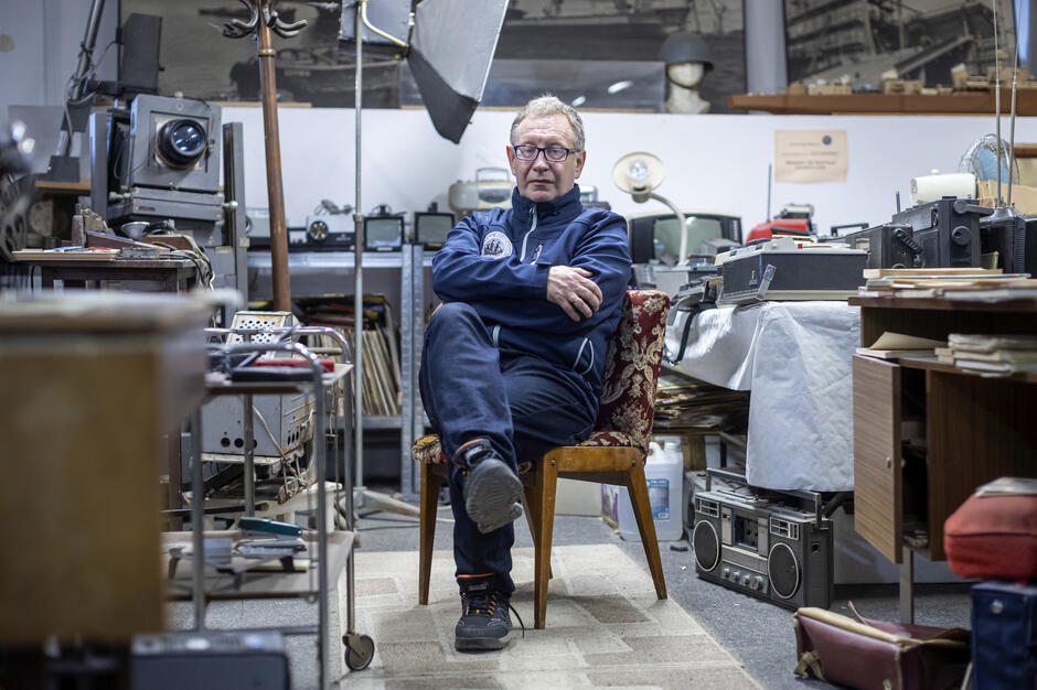 Mężczyzna siedzi na krześle z założonymi rękami i nogą, w otoczeniu licznych starych urządzeń technicznych, narzędzi oraz sprzętu fotograficznego. Za nim widać półki z archiwalnymi przedmiotami i duże zdjęcia przedstawiające stoczniowe sceny.
