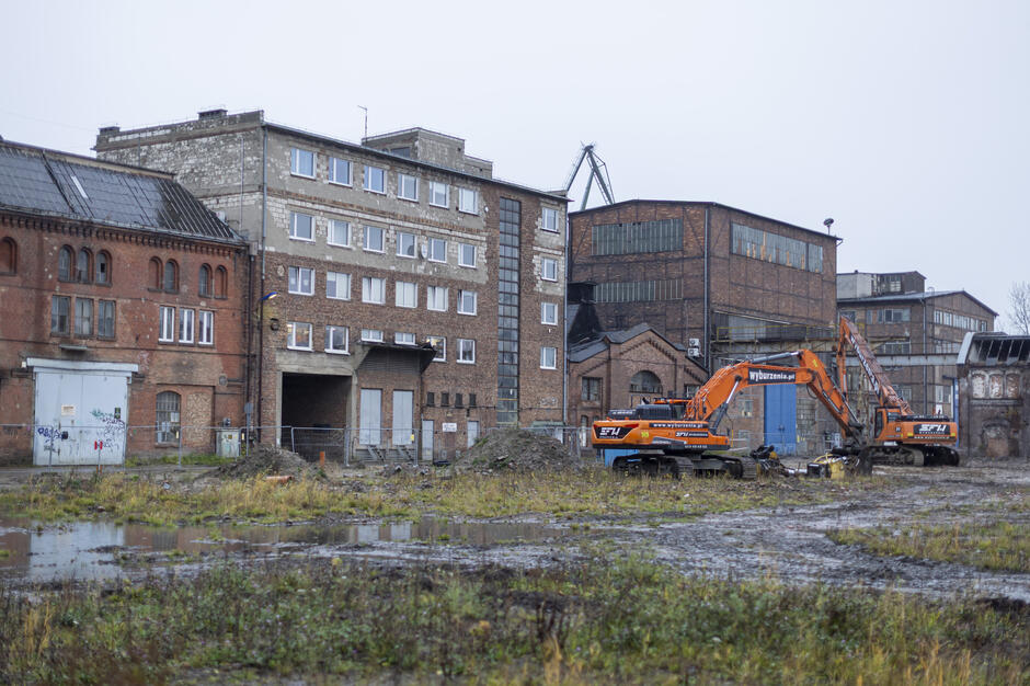 Na pierwszym planie widać błotnisty plac budowy z kałużami, na którym stoją dwie koparki w pomarańczowych barwach. W tle znajdują się stare, ceglane i betonowe zabudowania przemysłowe z dużymi oknami, noszące ślady zużycia i przygotowań do prac rozbiórkowych.
