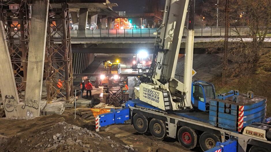 Na zdjęciu widać pracowników budowlanych w odblaskowych kamizelkach, którzy prowadzą nocne roboty pod konstrukcją rozbieranego wiaduktu, otoczeni rusztowaniami i ciężkim sprzętem. W tle działają maszyny o silnym oświetleniu roboczym, a teren wokół jest pełen ziemi, drewna i elementów metalowych wspierających konstrukcję.
