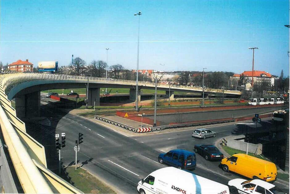 Widok na estakadę biegnącą łukiem nad ruchliwym skrzyżowaniem, otoczoną zabudową miejską i torami tramwajowymi. Na dole widać samochody czekające na światłach, a w tle jasne, słoneczne niebo.