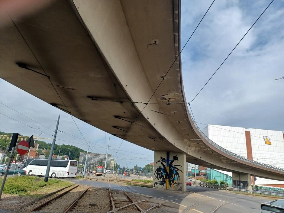 Ujęcie spod estakady ukazujące surowy beton i instalacje pod konstrukcją, z torami tramwajowymi i ulicą rozchodzącymi się w różnych kierunkach. Na jednym z filarów widoczny jest kolorowy mural przedstawiający abstrakcyjną postać.