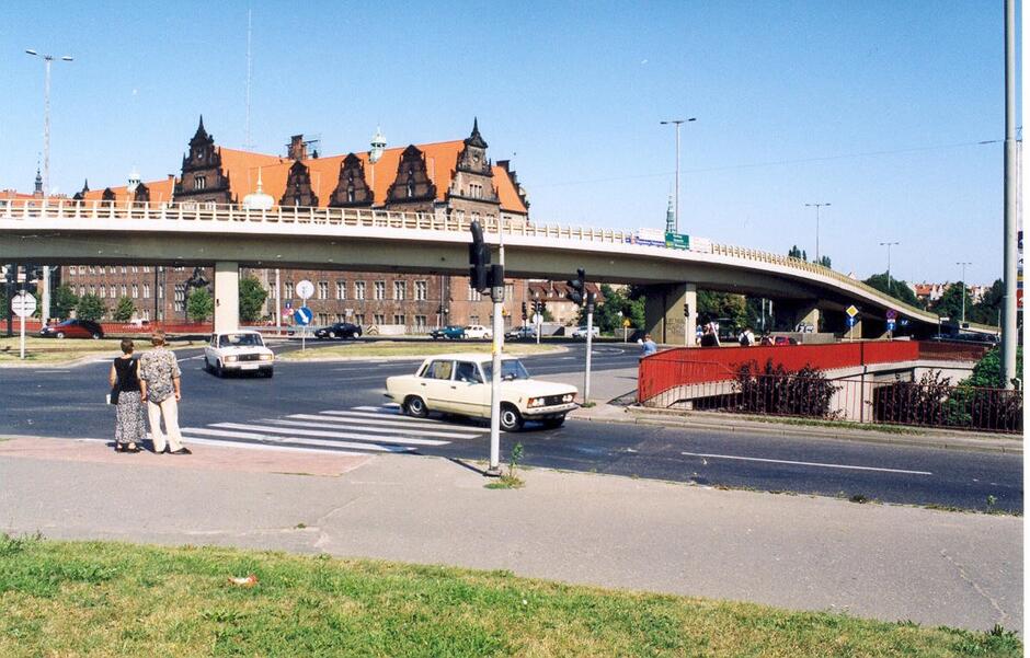 Estakada rozpięta nad szerokim skrzyżowaniem, na którego tle stoi zabytkowy gmach z czerwonym dachem i dekoracyjnymi szczytami. Przechodnie i samochody poruszają się po przejściach i ulicach pod konstrukcją