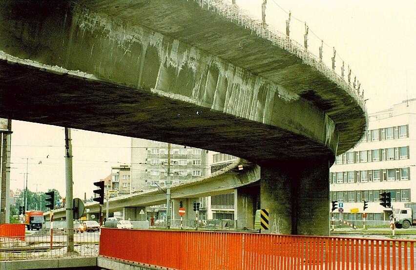 Bliski kadr uszkodzonej estakady, pokazujący popękany beton i widoczne wzmocnienia konstrukcyjne. Poniżej biegną tory tramwajowe i fragment czerwonej balustrady wejścia do przejścia podziemnego.