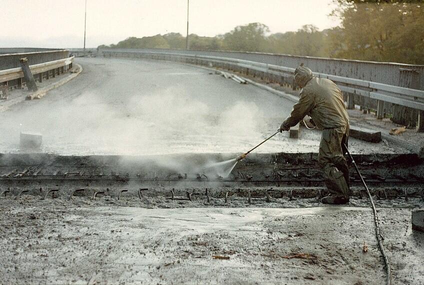 Pracownik w kombinezonie usuwa nawierzchnię estakady strumieniem wody pod wysokim ciśnieniem, a spod warstw asfaltu wyłaniają się metalowe pręty. Powierzchnia jest mokra, pełna błota i pary unoszącej się nad miejscem pracy.