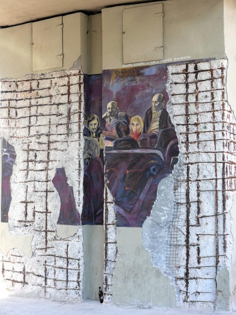 Filary estakady pokryte muralem przedstawiającym grupę ludzi, częściowo odsłoniętym przez usunięty tynk ukazujący zbrojenie betonu. Zniszczenia kontrastują z intensywnymi kolorami obrazu.