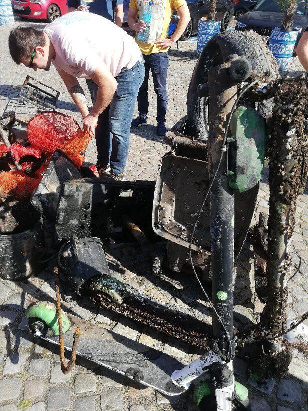 Kilka osób przegląda stertę odpadów wyciągniętych z wody, wśród których znajdują się zardzewiałe hulajnogi, metalowe elementy i plastikowe pojemniki. Widok ukazuje skalę zanieczyszczeń oraz wysiłek włożony w oczyszczanie przestrzeni publicznej.
