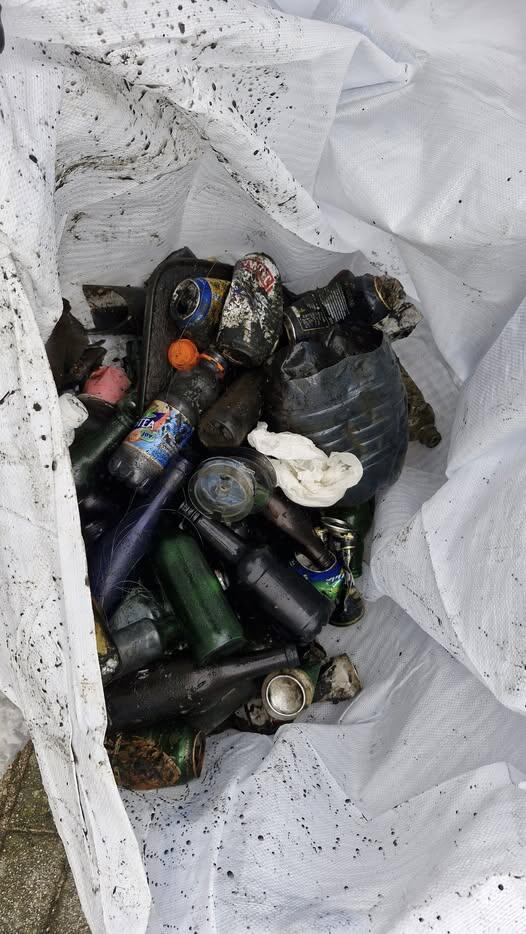 W dużym białym worku znajdują się liczne butelki, puszki i plastikowe opakowania zabrudzone mułem i rdzą. Zebrane odpady pokazują efekty sprzątania terenu lub zbiórki śmieci z wody.
